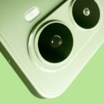 The Mivi One 5G smartphone in a matte Sage Green finish resting on a modern wooden desk beside wireless earbuds.