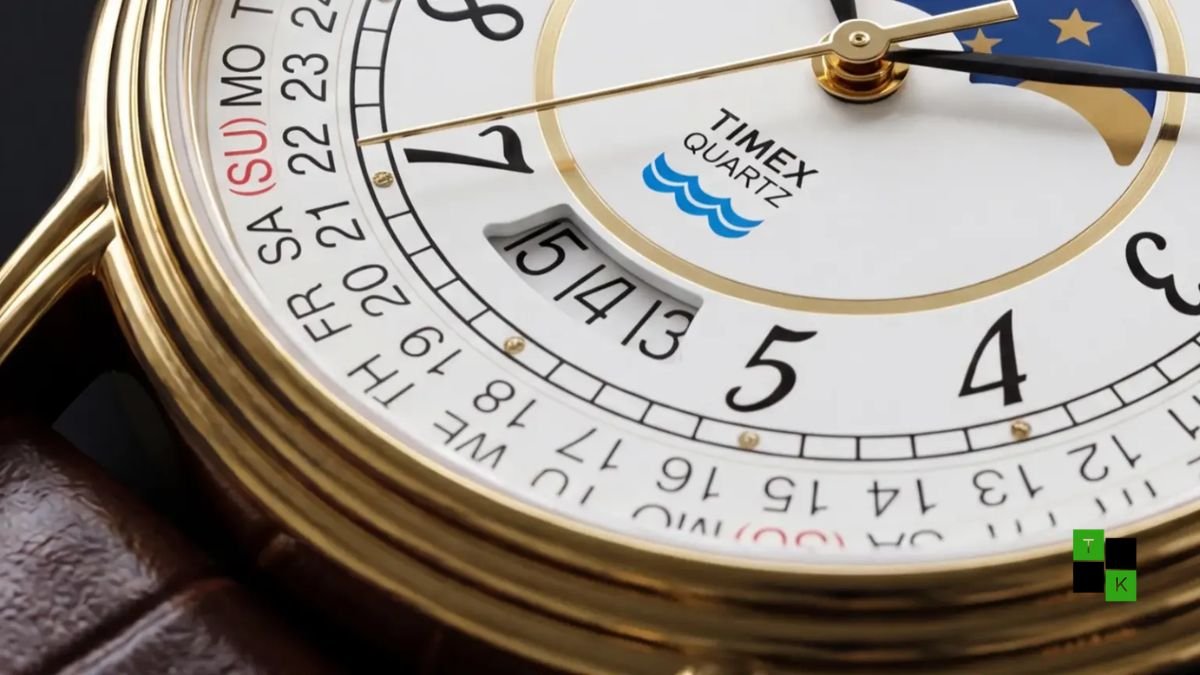 The Timex 1990 Moon Phase and Calendar Dial Reissue watch in gold-tone steel with brown leather strap resting on a dark walnut table.