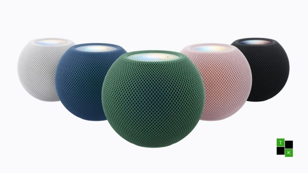 Five Apple HomePod mini 2 smart speakers in Midnight, White, Blue, Pink, and Green arranged in a semicircle on a sleek wooden table with glowing Sound Glow tops.